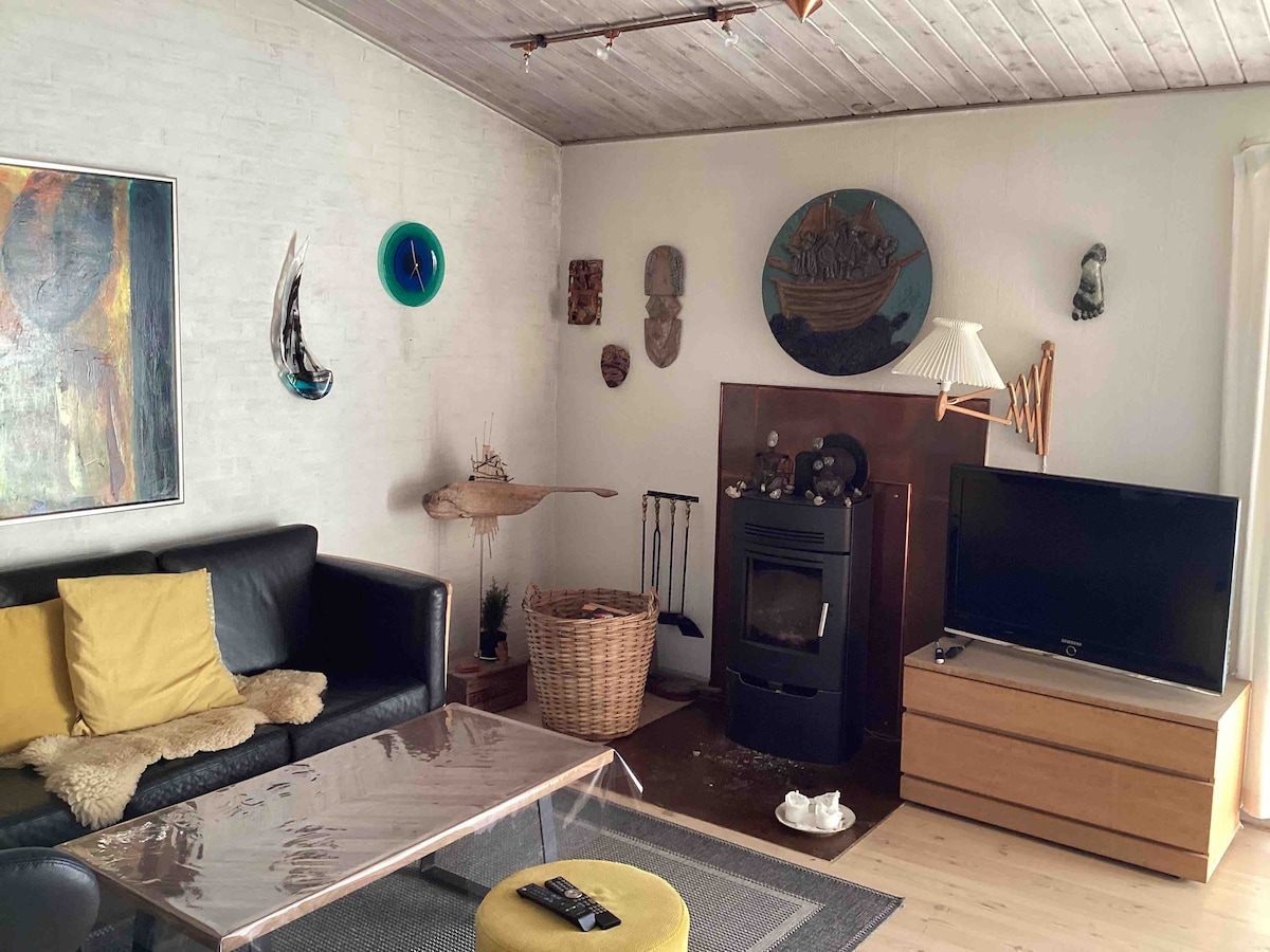 A cozy living area features a black leather sofa adorned with yellow cushions. A glass coffee table rests on a textured rug, while a television is positioned on a wooden media unit. Various artworks and decorative items decorate the walls and nearby surfaces.
