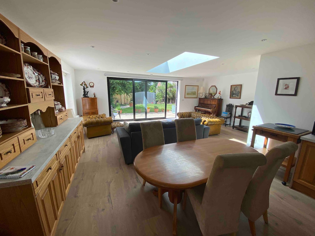 An open-plan kitchen, dining, and lounge area is illuminated by natural light. A wooden dining table is surrounded by upholstered chairs. A comfortable sofa is positioned near a large glass door that opens to a garden, with a skylight enhancing the bright atmosphere.