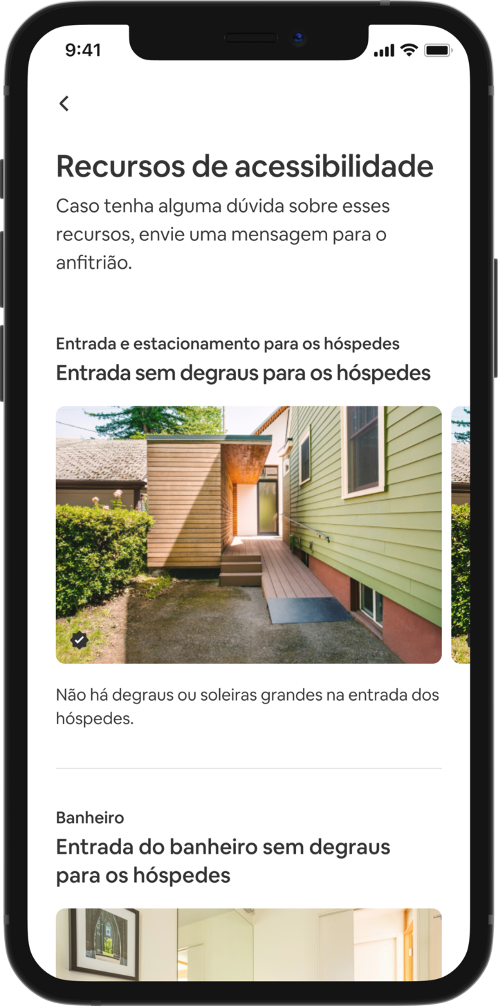 Um telefone celular exibe as informações de um anúncio do Airbnb. O título principal diz "Recursos de acessibilidade" e os recursos são agrupados por área, como "Entrada e estacionamento para hóspedes" e "Banheiro". Abaixo de "Entrada e estacionamento para hóspedes" está a informação "Entrada para hóspedes sem degraus" e a descrição da característica dizendo que "Não há degraus ou soleiras grandes na entrada para hóspedes." Abaixo está um carrossel de imagens com fotos dos recursos da acomodação, começando com uma rampa para cadeira de rodas.