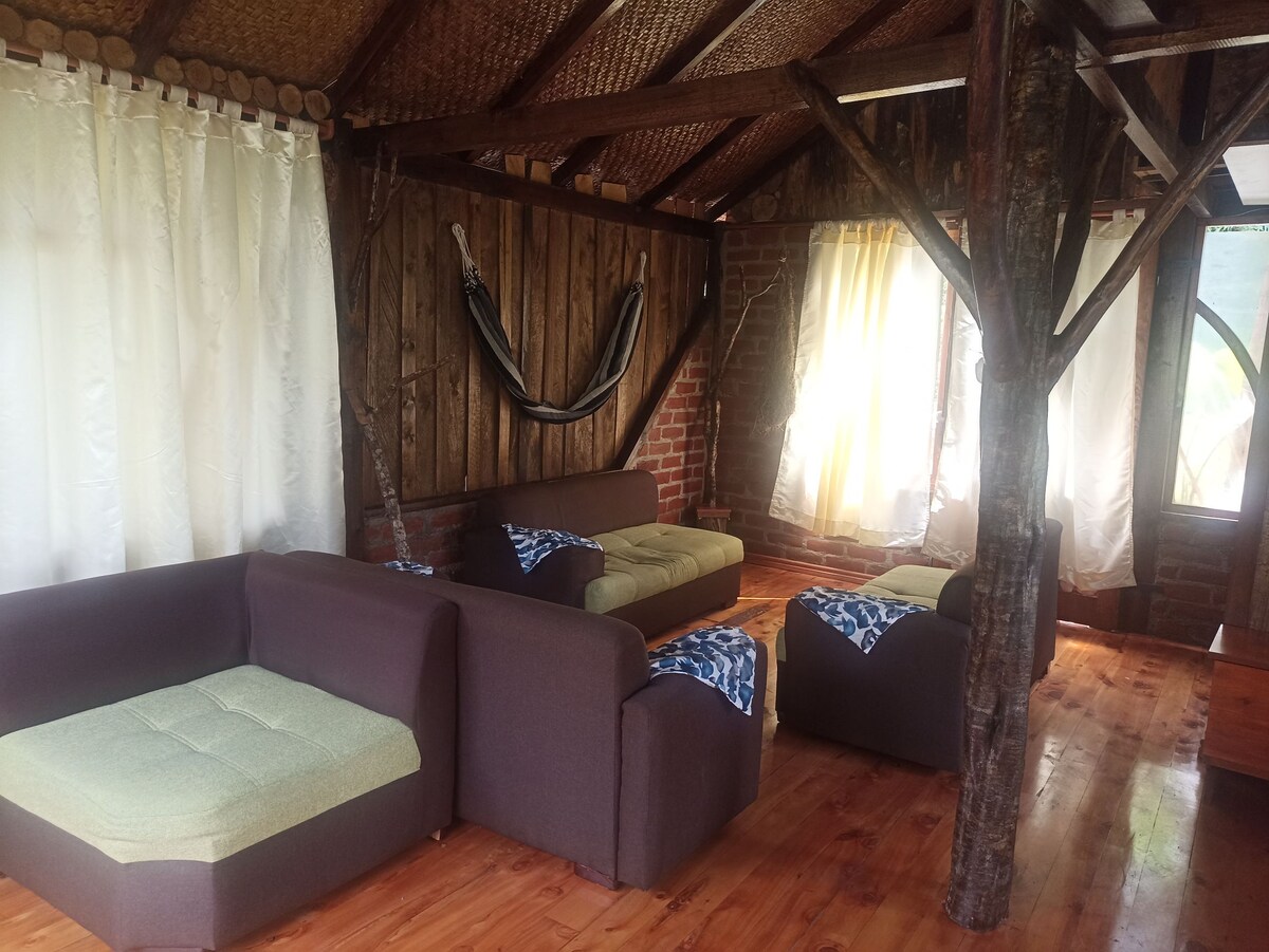 A cozy living area features earthy wooden beams and exposed brick walls. Soft seating in neutral tones surrounds the space, with blue and white throws draped casually over them. A hammock is suspended nearby, and light filters through sheer curtains, creating a relaxed atmosphere.