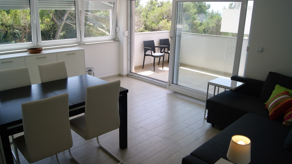 A bright living space features a dining table with white chairs and a sleek black table. Large glass doors lead to a private balcony, which contains two outdoor chairs. Natural light fills the area, highlighting light-colored tile flooring and modern furnishings.