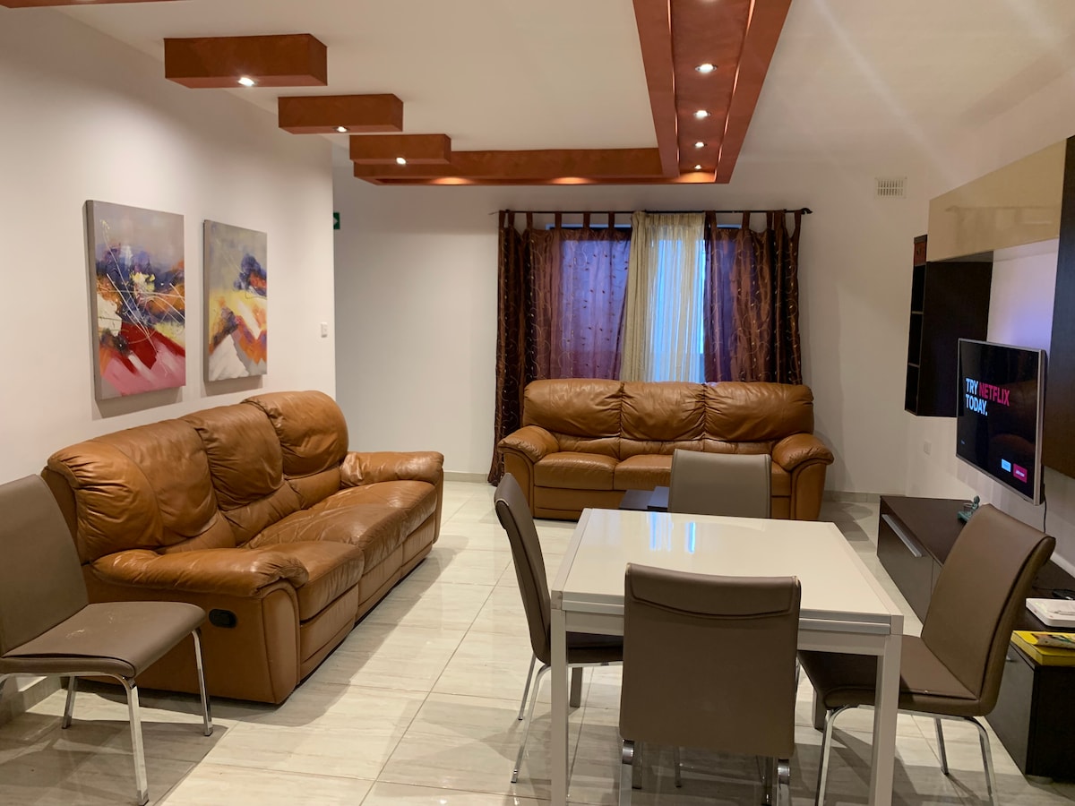 A spacious living area features two leather sofas and a modern dining table surrounded by chairs. Natural light enters through a window dressed with dark curtains. Colorful artwork adorns the walls, and a large flat-screen television is positioned against the opposite wall.