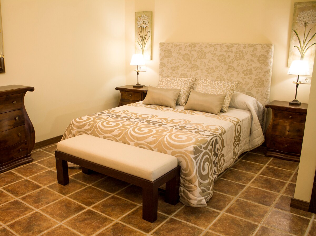 A comfortably furnished bedroom features a patterned bedspread draped over a spacious bed. Two bedside tables with lamps are positioned beside the bed. A wooden bench is centered at the foot of the bed, and decorative wall art adds subtle elegance to the room.