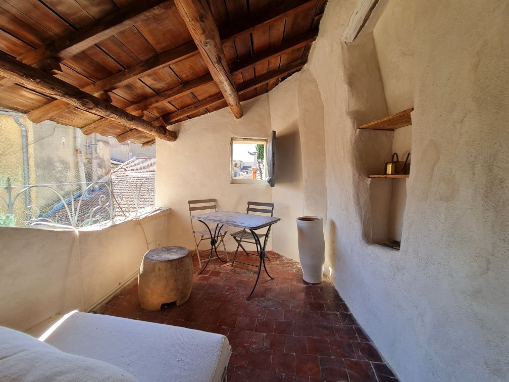 A private terrace features a rustic design with exposed wooden beams and terracotta flooring. A small table with two chairs sits against the wall, while a decorative vase and a natural stool add visual interest to the space. A window provides a view of the surroundings.