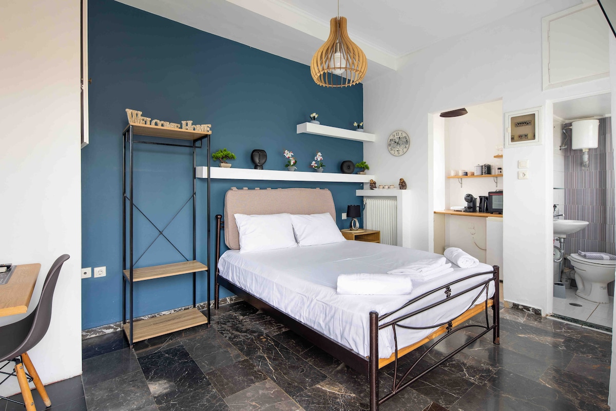 A comfortable sleeping area is showcased with a spacious bed dressed in white linens. A deep blue wall provides a backdrop, while floating shelves display decorative items. Dark flooring complements the modern furniture, and a kitchenette is visible in the corner.