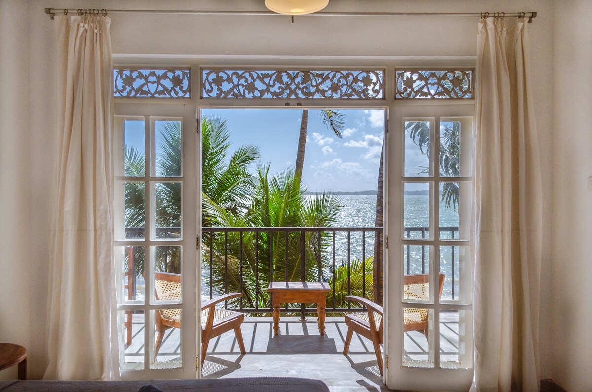 A private balcony is viewed through glass doors, showcasing two wicker chairs and a small table. Lush palm trees frame the ocean scene, with gentle waves visible in the distance under a clear blue sky.