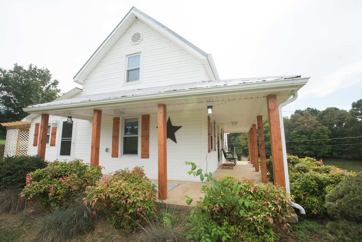 The exterior of the cottage is shown, highlighting its white siding and prominent star decoration. Wooden pillars offer support to the porch overhang, while lush greenery and landscaping surround the foundation, providing a welcoming and serene atmosphere.