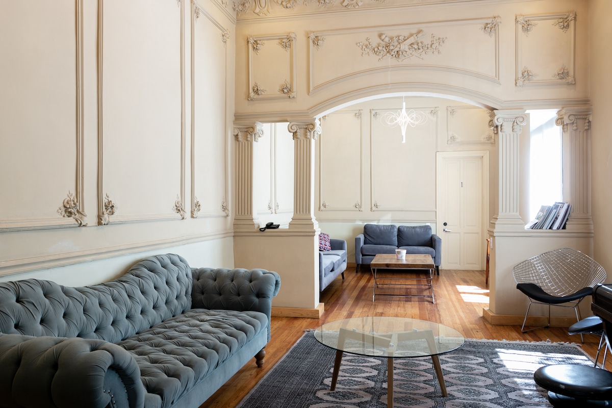 A spacious living area displays ornate wall details and high ceilings. A light-colored coffee table sits at the center, surrounded by a plush, tufted sofa and additional seating. Natural light flows through large windows, creating a bright and inviting space.