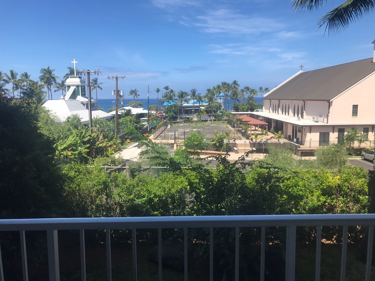 A view from the lanai features lush green vegetation in the foreground with a glimpse of the ocean in the background. The landscape is dotted with palm trees and includes nearby buildings, showcasing a peaceful setting.