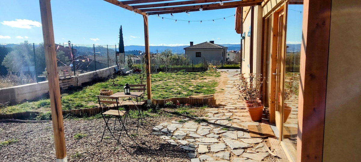 A stone pathway leads to a spacious outdoor area, featuring a wooden pergola supported by rustic pillars. A small table and two chairs are arranged nearby, with lush greenery and distant hills visible in the background, under a clear blue sky.