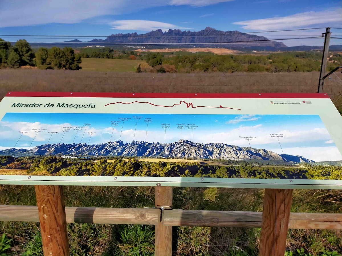 A viewpoint sign is displayed prominently, featuring a panoramic illustration of Montserrat's mountain range. The surrounding landscape includes rolling hills with greenery, and the mountains rise impressively in the background, showcasing their distinct rocky formations. Clear skies enhance the natural beauty of the scene.