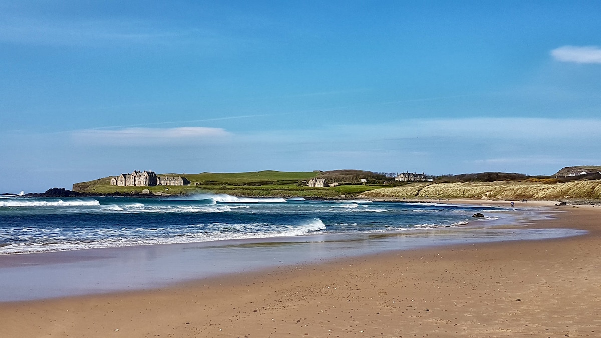 A sandy beach is presented with gentle waves lapping at the shore. In the distance, grassy hills rise, dotted with a few historical buildings. The sky is clear with soft clouds, creating a serene atmosphere that enhances the coastal landscape.