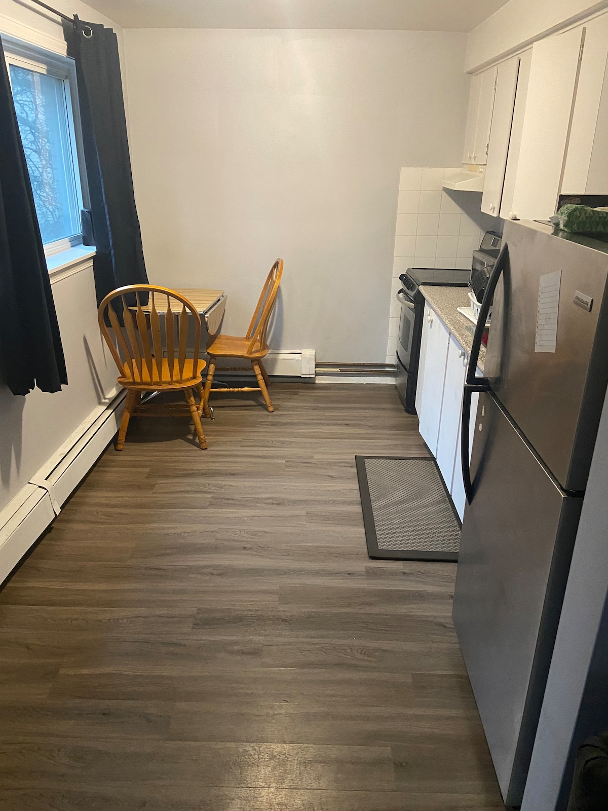 A clean and functional kitchen area is visible, featuring a stainless steel refrigerator and a microwave. Two wooden chairs are paired with a small table against the wall, complemented by white cabinets and a tiled backsplash. Natural light enters through a nearby window, framed by black curtains.