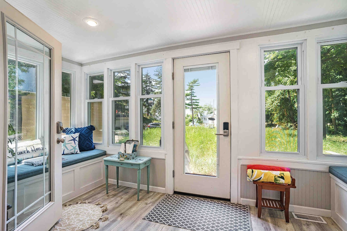 A sunlit porch features large windows allowing natural light to fill the space. A small blue table is positioned near the entrance, flanked by two cushioned benches. A door opens to a view of greenery outside, creating a serene connection with the outdoors.