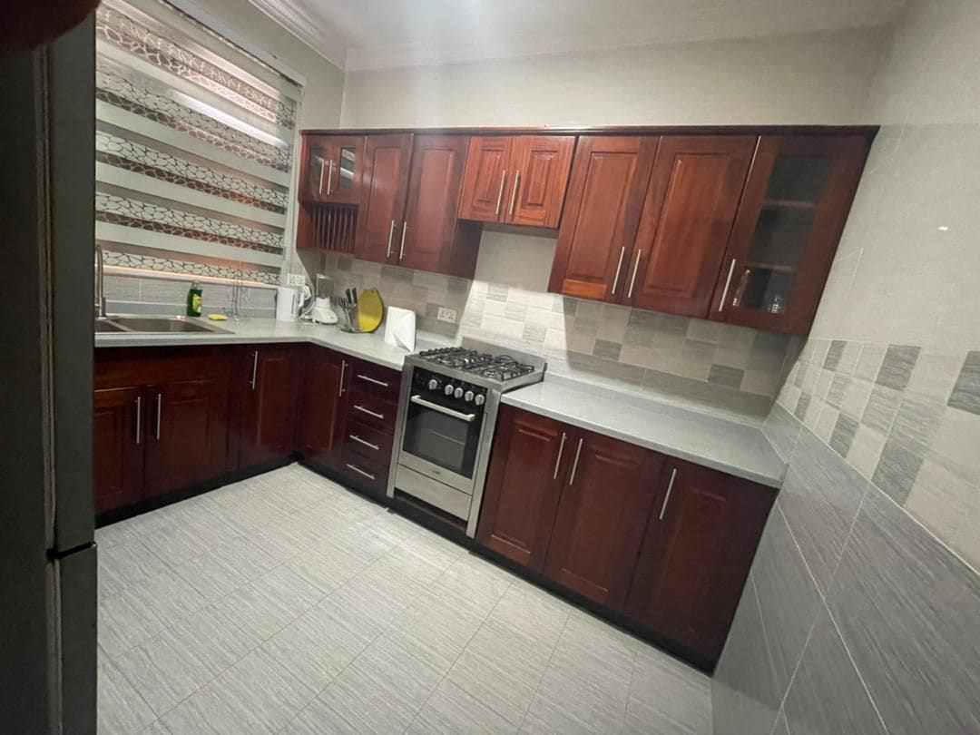 The kitchen features modern wooden cabinetry with a sleek finish and a spacious countertop. Stainless steel appliances, including a gas stove and oven, are present. A window with patterned shades allows natural light to illuminate the space, enhancing functionality.