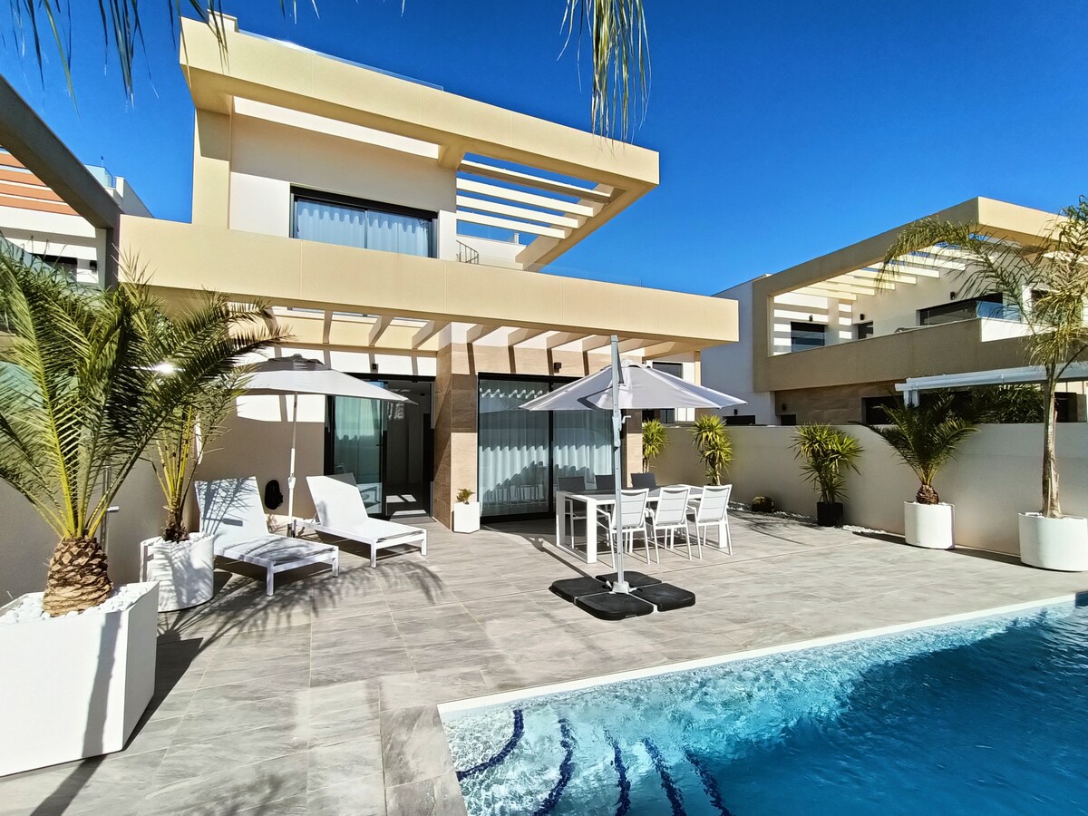 A modern villa features a spacious outdoor area with a private swimming pool and sun loungers. Two umbrellas provide shade over a dining table set for six. Palm trees are planted in white planters, enhancing the contemporary design against a clear blue sky.