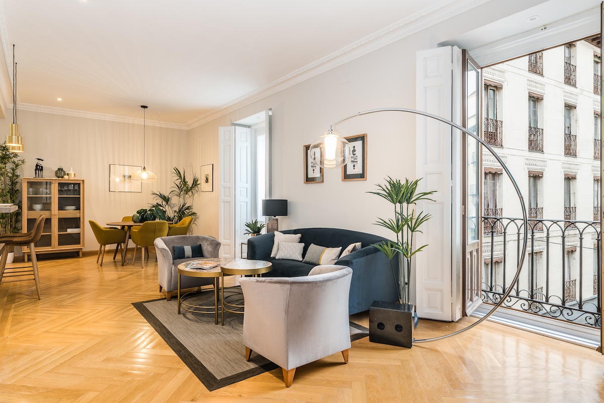 A spacious living room is characterized by a modern design with a large blue sofa and two plush armchairs. A pair of round coffee tables is set on a neutral rug, surrounded by decorative plants. Natural light fills the room through two open balcony doors, revealing views of the building's exterior.