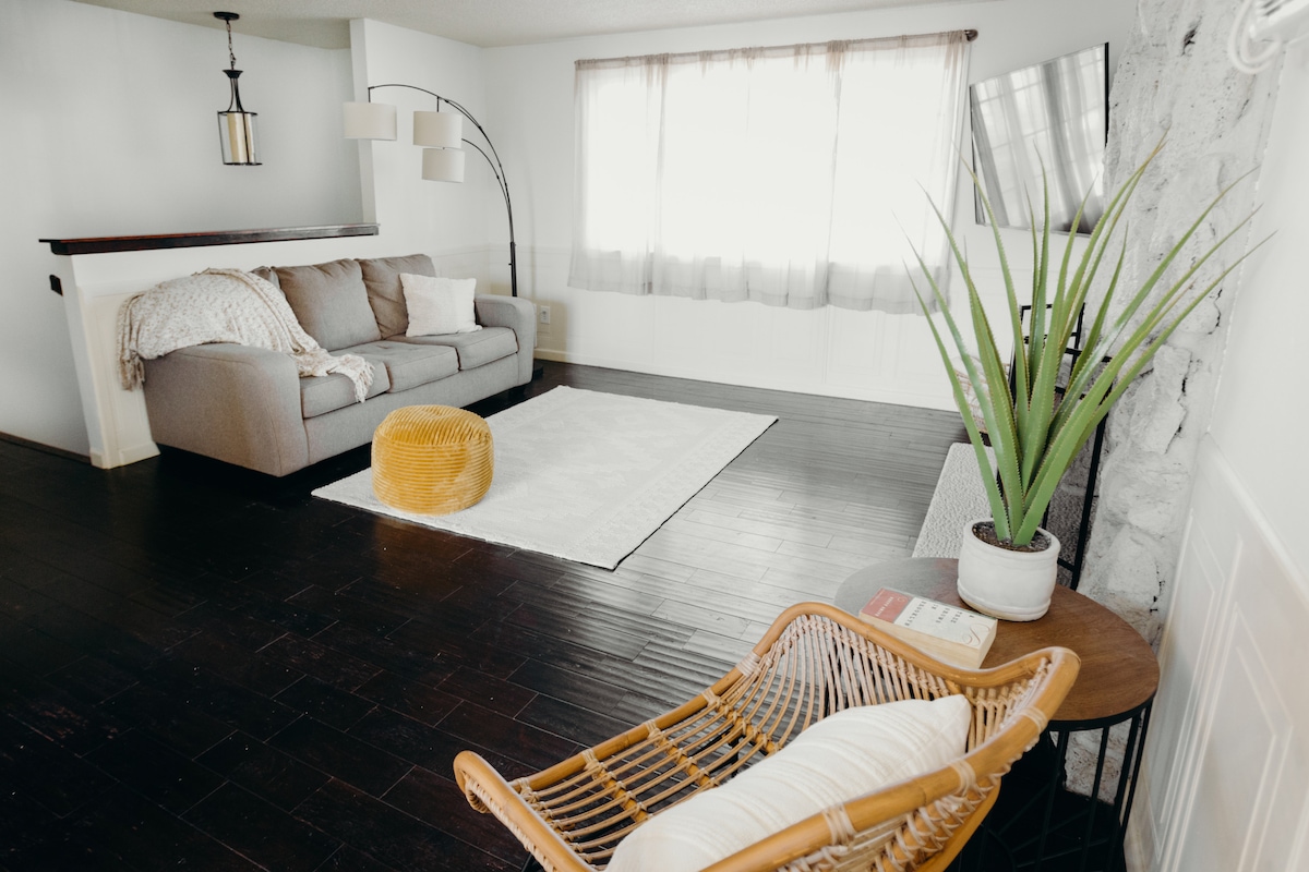 A spacious living area is highlighted by a comfortable gray sofa, a decorative yellow pouf, and a stylish rattan chair beside a small side table. Large windows provide natural light, while a simple area rug adds warmth to the dark wooden flooring.