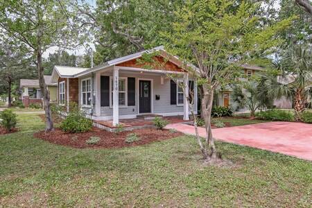 A charming cottage with a welcoming front porch is surrounded by well-maintained landscaping. Lush greenery and flowering plants enhance the inviting exterior. The driveway provides ample parking space, and a tree offers shade in the front yard for a relaxed outdoor atmosphere.