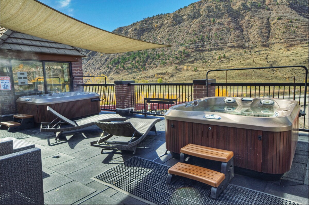 An outdoor hot tub area is showcased with two inviting hot tubs, surrounded by a landscape of mountains. Comfortable lounge chairs are positioned for relaxation, and shaded sections provide respite from sunlight, enhancing the area's appeal for unwinding after a day of exploration.