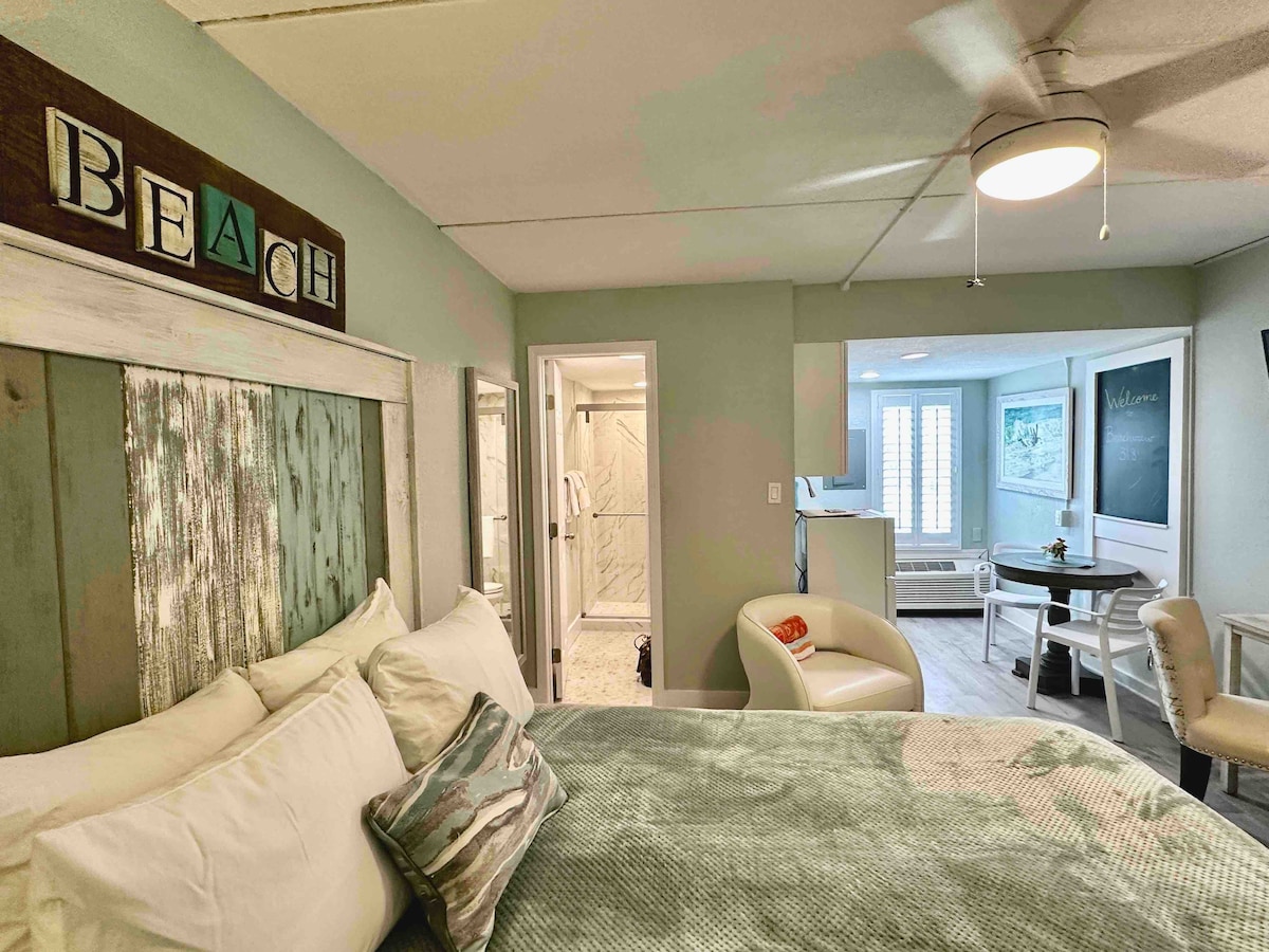 A spacious studio features a large bed with decorative pillows and a soft green throw. An ensuite bathroom is visible through an open door, along with a white armchair and a small dining table in the background. Natural light filters through the window, illuminating the welcoming space.