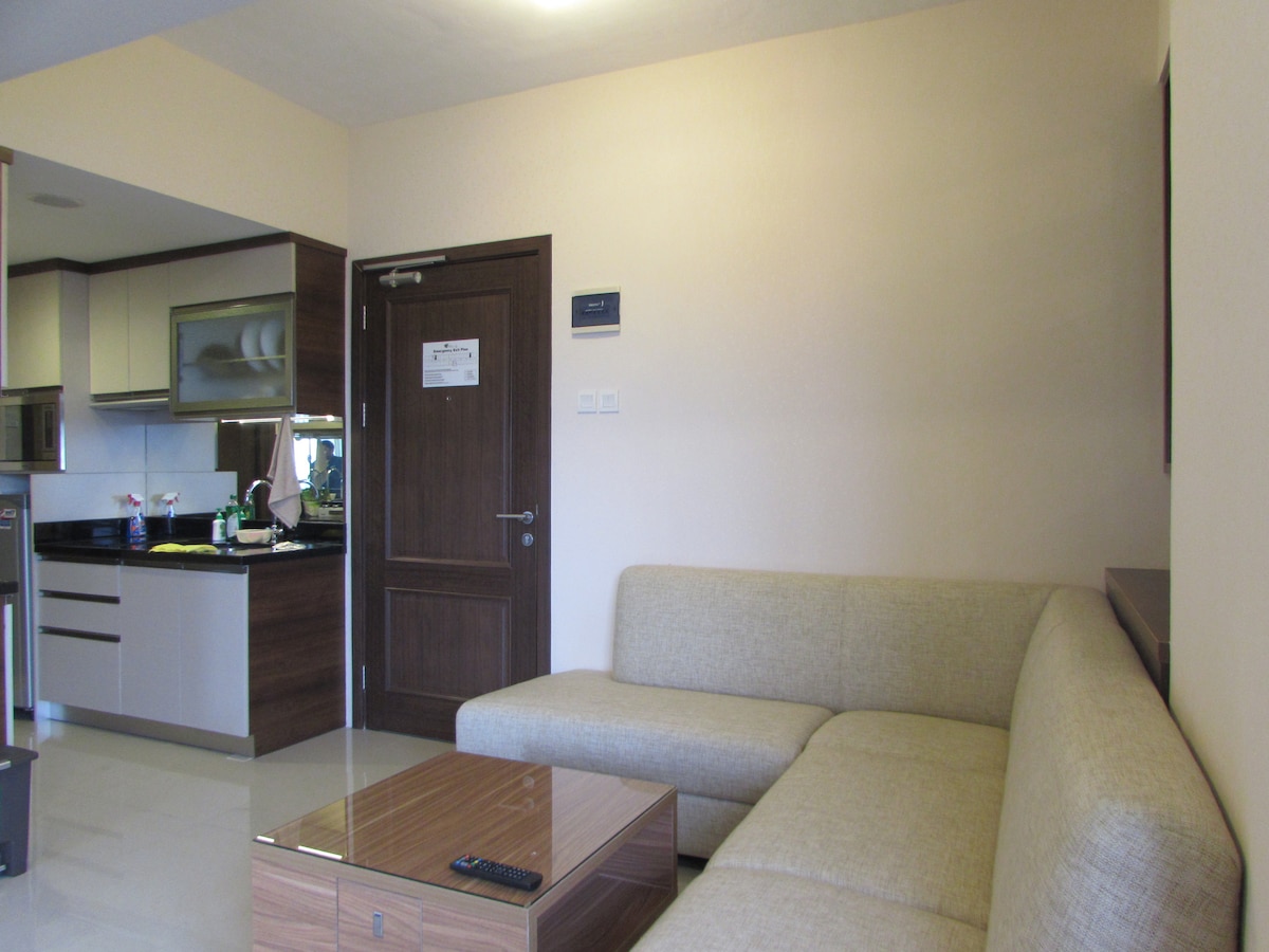 A spacious living area showcases a comfortable L-shaped sofa facing a low coffee table. A doorway leads to another room, while a modern kitchen is visible in the background, featuring sleek cabinetry and essential appliances, highlighting the open layout of the apartment.