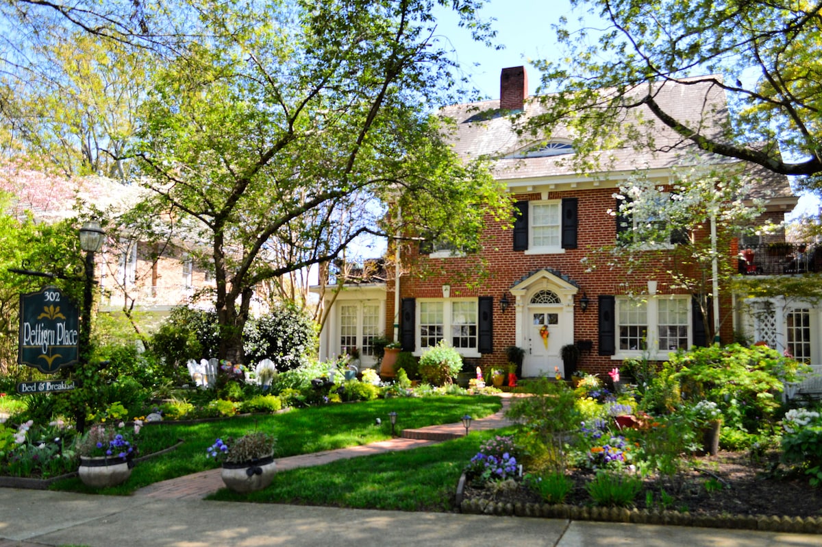 A red brick Federal-style home is surrounded by a well-tended garden featuring blooming flowers and lush greenery. Tall trees provide shade over the manicured lawn, while the inviting front porch showcases classic architectural details inviting guests to explore.