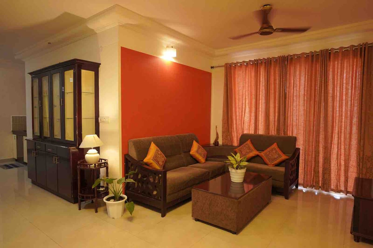 A comfortable living area features a pair of sofas adorned with orange cushions. A coffee table sits centrally, while a potted plant adds a touch of greenery. Large windows, dressed with light-filtering curtains, provide ample natural light, enhancing the inviting atmosphere.