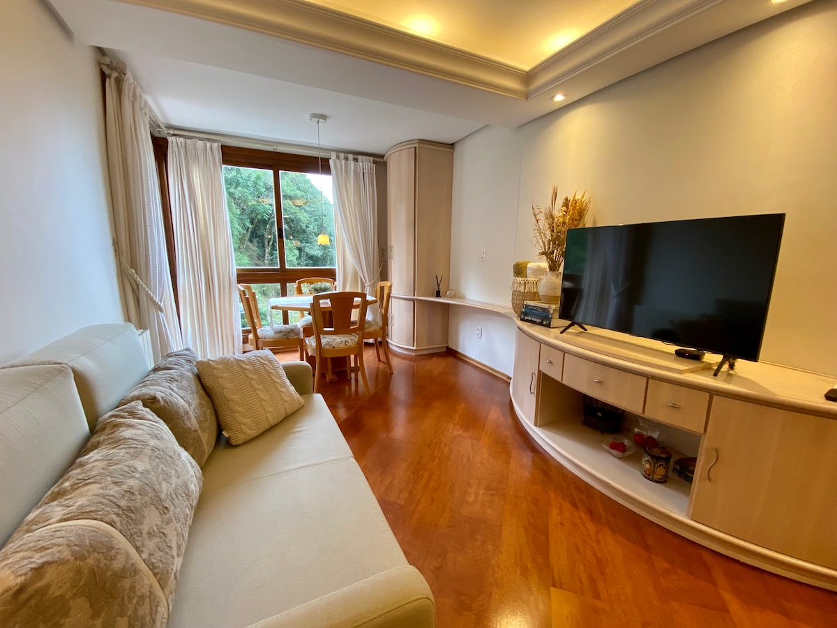 A living area features a light-colored sofa paired with a decorative pillow, complemented by a wooden entertainment unit housing a large Smart TV. Natural light fills the space through a large window, which is framed by sheer white curtains, revealing a view of greenery beyond.