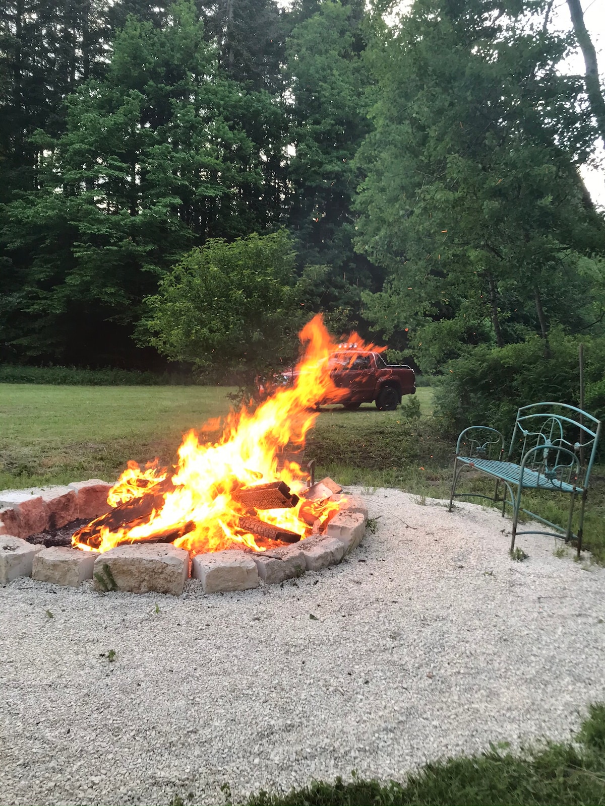 A stone fire pit is surrounded by gravel, with vibrant flames rising from a wood fire. Green trees and a grassy area provide a natural backdrop. A metal chair is positioned near the fire pit, inviting relaxation. A vehicle is partially visible in the background.