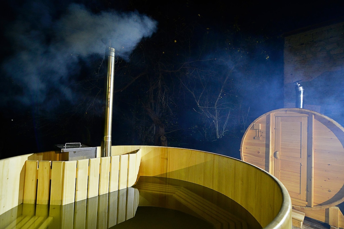 A traditional wooden hot tub is surrounded by swirling steam, creating a serene atmosphere. A second wooden structure, resembling a sauna, is positioned nearby. The setting is illuminated by soft lighting against a backdrop of trees.