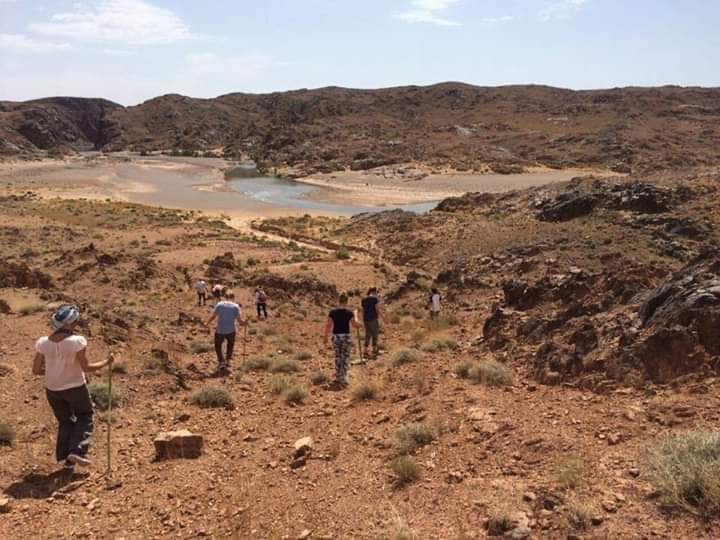 A group of individuals is seen hiking down a rocky terrain, surrounded by arid landscapes. A river is visible in the background, winding through the valley. The sky is clear with a few clouds, casting soft shadows across the ground.