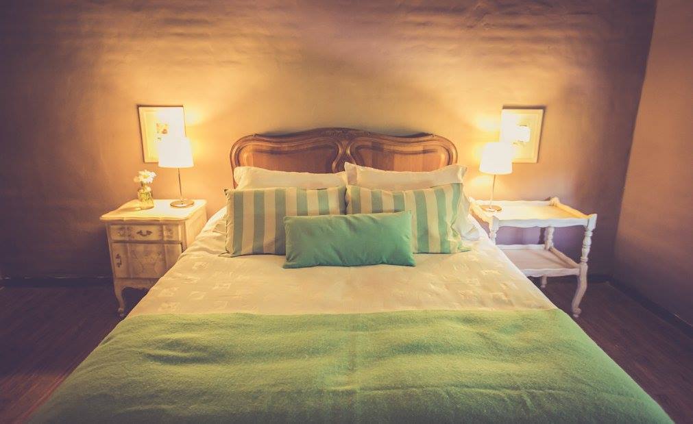 A cozy bedroom is presented with a spacious bed layered with white linens and soft green accents. Two bedside tables with lamps flank the bed, and framed artwork is visible on the walls, creating a serene and inviting setting.