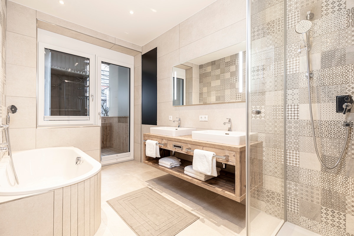 The spacious bathroom features a large freestanding bathtub alongside a wooden vanity with two sinks. A walk-in shower with contemporary tiles is visible. Natural light enters through a window, illuminating the clean, neutral tones of the design.