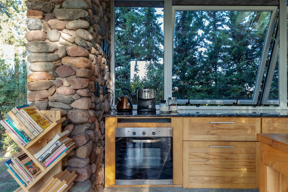 A kitchen area features a combination of wooden cabinetry and stone walls. A modern stove is positioned beneath a countertop, with a kettle and coffee maker nearby. A bookshelf displays a variety of books, and large windows provide views of the lush greenery outside.