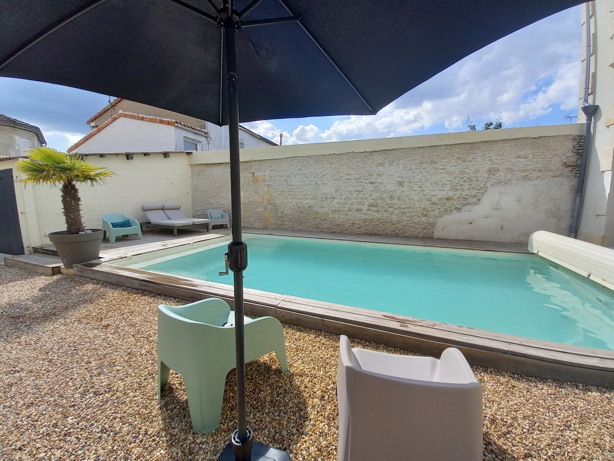 The image captures a serene swimming pool enclosed within a stone wall, featuring a wooden deck. Lounge chairs are positioned around the pool, while an umbrella provides shade. A small palm tree adds a touch of greenery to the gravel area surrounding the pool.