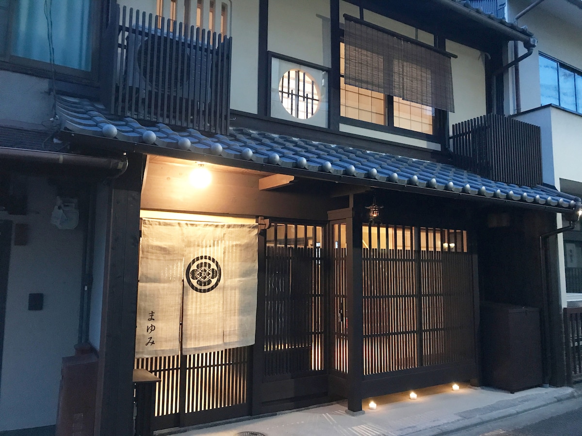 The exterior of a traditional Japanese townhouse features a wooden facade with a tiled roof. Soft lighting bathes the entrance, which is adorned with a decorative curtain. Windows display unique shapes and patterns, contributing to the charm of this welcoming space.