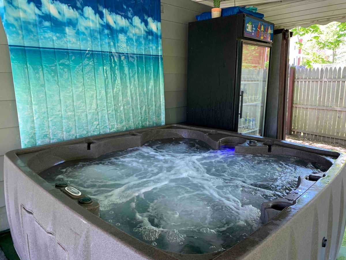 A hot tub is seen bubbling, set against a backdrop of a beach-themed curtain depicting a serene ocean scene. A dark cabinet is positioned nearby, providing storage space. The surrounding area is well-lit and features inviting green flooring.