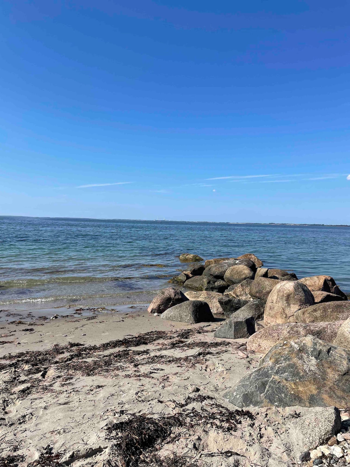 A sandy beach extends towards clear waters, where a rocky outcrop is visible along the shoreline. Seaweed lies scattered across the shore, and the sky is bright blue with a few wispy clouds. A calm and tranquil atmosphere is suggested by the serene setting.