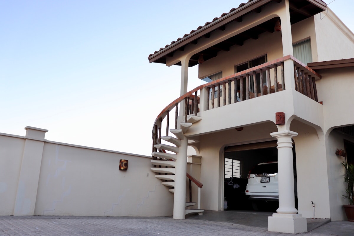 A two-story home is showcased, featuring a distinctive spiral staircase leading to an upper balcony. The automobile is parked in the garage beneath, with a smooth driveway extending before the residence and a beige wall enclosing the property.