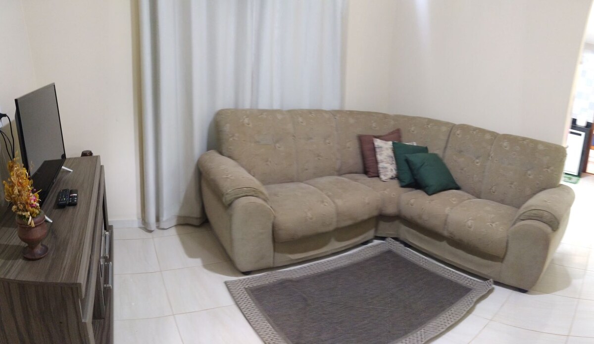 A comfortable seating area features a spacious light brown sectional sofa adorned with decorative pillows. A small area rug lies beneath the sofa, and a television is positioned on a wooden stand nearby. Natural light filters through a sheer curtain.