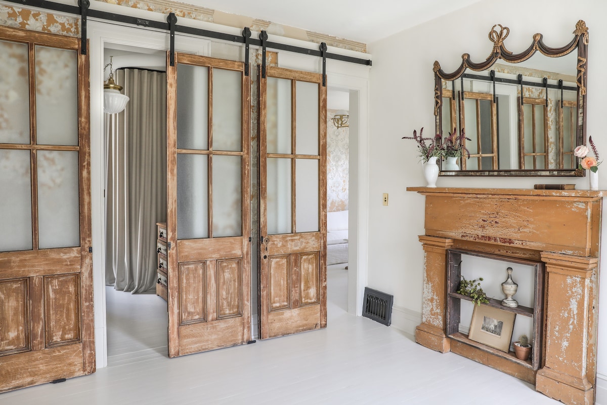 A warm entryway features sliding wooden doors with glass panels, leading into a spacious room. An ornate mirror hangs above a vintage fireplace, complemented by decorative items. Soft natural light enhances the wood tones and neutral colors of the walls.