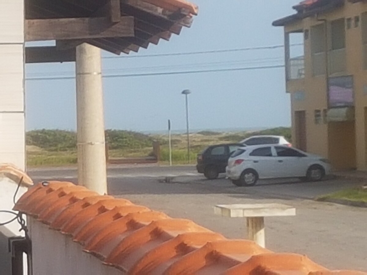 A view is provided from the property, showcasing a glimpse of the beach in the distance. Two parked cars are visible on the paved street, with a street lamp positioned nearby. Soft greenery partly frames the background, adding to the outdoor scene.