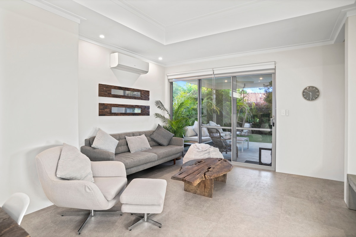 A modern living area is showcased, featuring a gray sofa with plush cushions, accompanied by a sleek armchair and ottoman. A wooden coffee table adds natural texture. Large sliding glass doors open to a lush outdoor space, allowing ample light to fill the room.