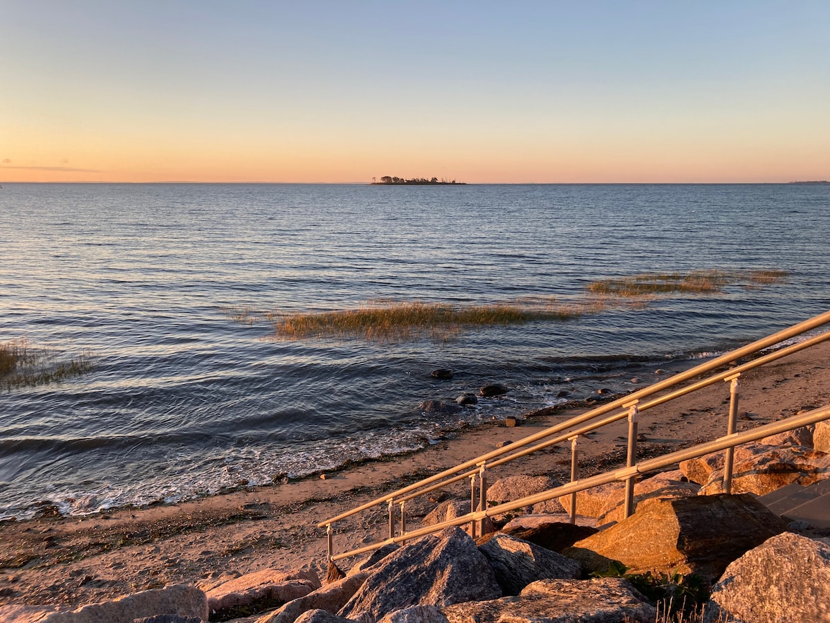 A serene view of the shoreline at dusk, showcasing gentle waves lapping against the sandy beach. A wooden railing leads down the steps to the water's edge, with smooth stones lining the path. A remote island can be seen in the distance, against a softly colored sky.