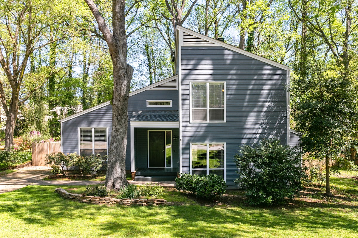 The exterior of the contemporary home showcases a unique angular architecture and large windows. A welcoming entrance is framed by a pathway leading to the door, while the surrounding lush greenery creates a peaceful, natural setting.