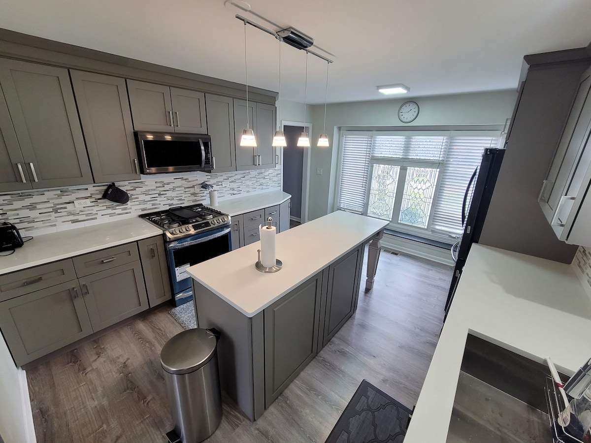 The kitchen features modern cabinetry in a soft grey tone, complemented by white countertops and a backsplash of patterned tiles. A large island with seating is positioned at its center, illuminated by pendant lights. A spacious window allows natural light, highlighting the clean, functional design.