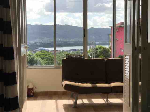 Large windows frame a stunning view of the mountains and sea, allowing natural light to fill the cozy seating area. A simple, modern couch is positioned to offer the best perspective of the landscape, enhancing the room's inviting ambiance.