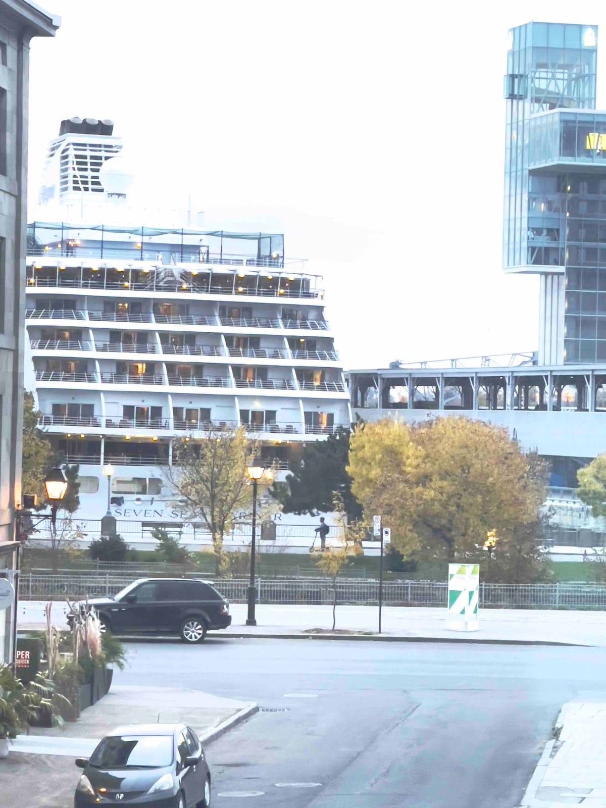 An overview of the waterfront is displayed, featuring a large cruise ship docked nearby. Lush trees with autumn foliage provide a splash of color, while modern buildings frame the scene. A street and parked vehicle are visible in the foreground.