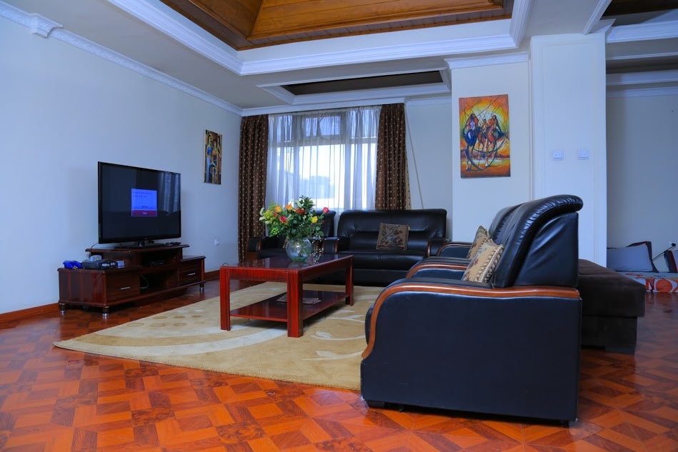 A spacious living area features a large flat-screen television, a dark leather sofa set, and a wooden coffee table. A plush area rug is positioned on the tiled floor, while natural light illuminates the room through large windows, complemented by patterned drapes.